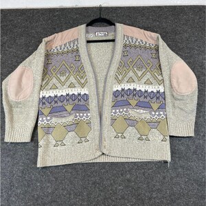 May include: A beige knit cardigan featuring a V-neckline, button-front closure, and long sleeves with tan elbow patches. The garment displays a multi-colored geometric pattern in shades of purple, olive green, and white, with tan shoulder panels.
