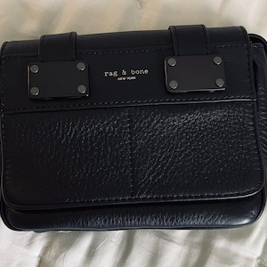May include: A navy blue leather crossbody bag with a flap closure. The bag features a textured surface, metal accents, and the text "rag & bone NEW YORK". The bag has a rectangular shape and a structured design.