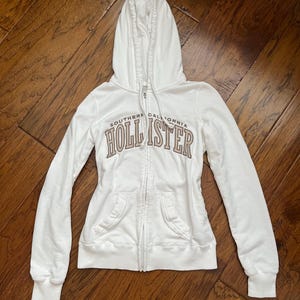 May include: White zip-up hoodie with a hood and front pockets. The hoodie features the text "SOUTHERN CALIFORNIA HOLLISTER" in brown lettering across the chest. The hoodie is made of a soft material.