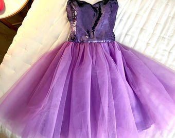 May include: A lavender and purple dress with a sequined bodice and a tulle skirt. The dress has thin black straps and is displayed on a white surface. The bodice is a darker shade of purple than the skirt.