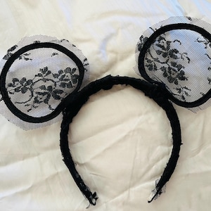 May include: Black lace mouse ear headband. The ears are circular with a black floral lace design over a white background. The headband is black and covered in a textured fabric.