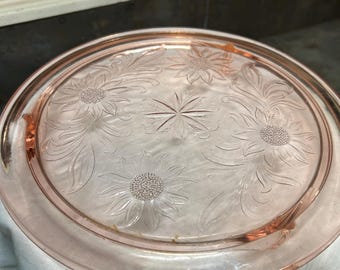 May include: A pink glass cake stand with a floral design. The stand features an embossed pattern of sunflowers and leaves. The edges are slightly curved, and the stand is resting on a white cloth.