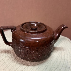 May include: A brown ceramic teapot with a textured lower half and a smooth, glossy upper half. The teapot has a handle, a spout, and a lid with a small knob. The teapot is designed for brewing tea and measures approximately 9 inches in length.