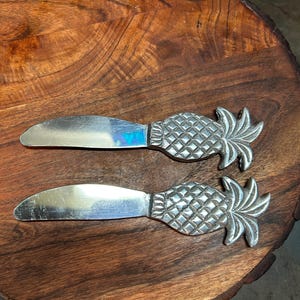 May include: Two silver butter knives with pineapple-shaped handles rest on a wooden cutting board. The knives have a shiny, reflective blade and a detailed pineapple design. The wooden board has a natural, rustic appearance.