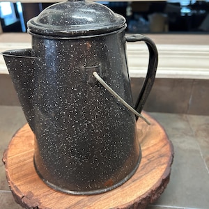 May include: A black and white speckled enamelware coffee pot with a lid and handle. The pot is sitting on a round, wooden slab with a natural edge. The pot has a spout for pouring.