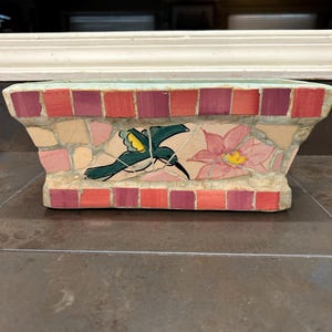 May include: A small, rectangular planter made of mosaic tiles. The planter features a pink flower and a green hummingbird with yellow accents. The planter is made of small, colorful tiles.