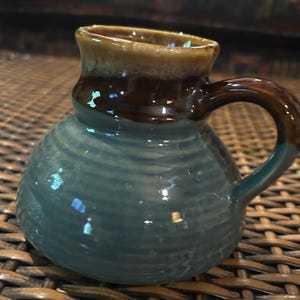 May include: A small ceramic mug with a rounded, bulbous body featuring a ribbed texture. The lower portion is a muted blue, while the upper neck and handle are finished in a glossy dark brown glaze with a lighter tan rim at the top opening.