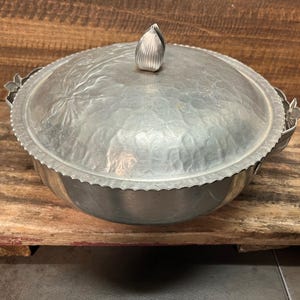 May include: A silver aluminum serving dish with a lid. The lid has a decorative design and a knob in the center. The dish has a scalloped edge.