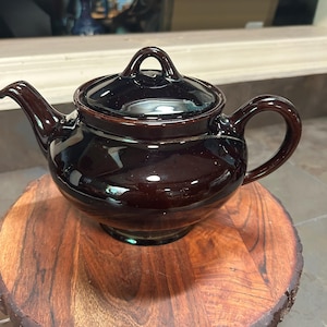 May include: A dark brown ceramic teapot with a matching lid and handle. The teapot is glossy and sits on a round wooden base. The spout and handle are curved, and the lid has a small loop for easy removal.