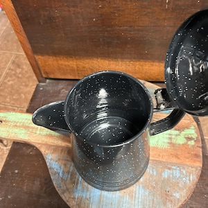 May include: A black enamel coffee pot with a speckled white pattern. The pot has a spout, handle, and a lid that is partially open. The pot is sitting on a wooden cutting board with blue and green paint.