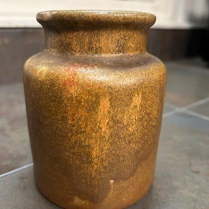 May include: A small, cylindrical ceramic vase with a textured, speckled surface. The vase has a warm, earthy color palette, with shades of brown and gold. The rim of the vase is slightly flared.