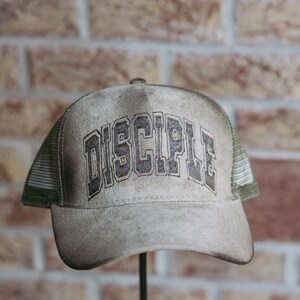 May include: A taupe-colored trucker hat featuring a mesh back and a solid front panel. The word "DISCIPLE" is printed in a dark, distressed block font across the center of the front panel. The hat has a curved brim and a structured crown design.
