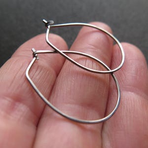 Peut inclure: Une paire de boucles d'oreilles argentées en forme de cerceau. Les boucles d'oreilles ont une forme de goutte d'eau avec un fond carré. Le métal est fin et semble léger. Les boucles d'oreilles sont présentées sur un fond neutre.