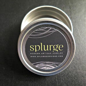 Puede incluir: Una lata redonda de plata con una etiqueta en blanco y negro que dice "splurge modern artisan jewelry www.splurgedesigns.com"