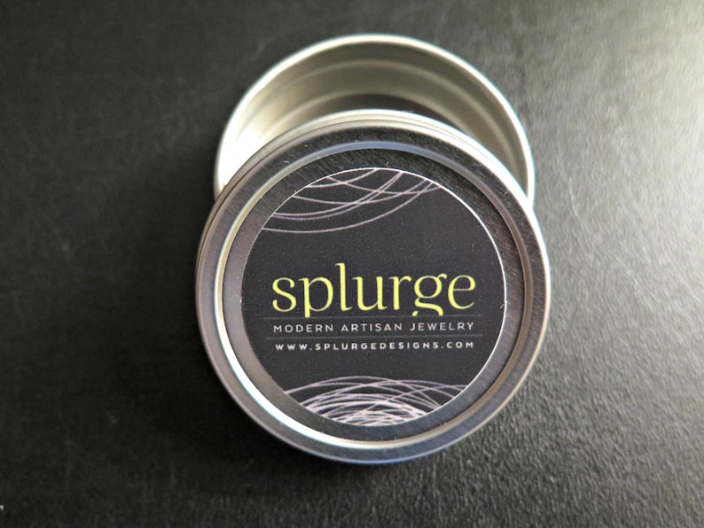 Puede incluir: Una lata redonda de plata con una etiqueta en blanco y negro que dice "splurge modern artisan jewelry www.splurgedesigns.com"