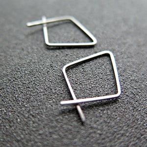 May include: A pair of silver-coloured, geometric square earrings. Each earring has a square shape with a post for insertion into the ear. The earrings are shown on a dark, textured surface.