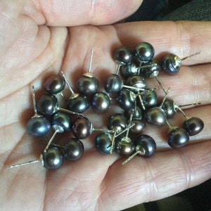 Puede incluir: Una colecci&oacute;n de pendientes de perlas gris oscuro con postes de plata. Las perlas son redondas y tienen un ligero brillo iridiscente.