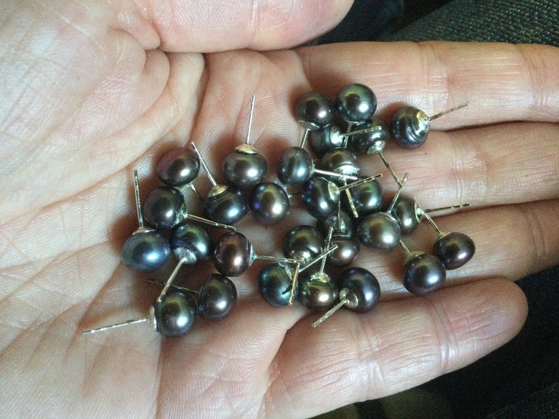 Puede incluir: Una colecci&oacute;n de pendientes de perlas gris oscuro con postes de plata. Las perlas son redondas y tienen un ligero brillo iridiscente.