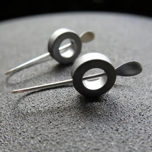 May include: A pair of silver earrings with a circular design. Each earring has a small, flat, teardrop-shaped piece attached to the side.