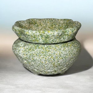 May include: Two-piece speckled stone bowl set. The bowls are light gray with green and yellow speckles. The top bowl has a scalloped edge and sits atop a larger, rounded bowl. The set is suitable for decorative or functional use.