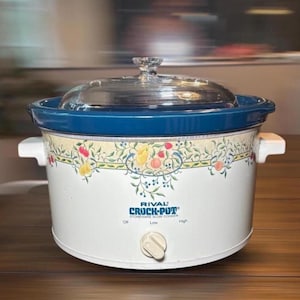 May include: A Rival Crock-Pot stoneware slow cooker with a white base, a blue rim, and a clear glass lid. The exterior features a decorative fruit and vine pattern. A single control knob is centered on the front with Off, Low, and High settings.