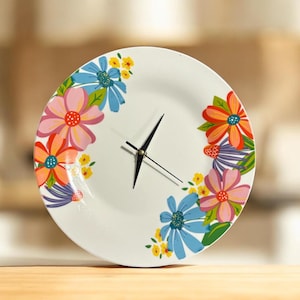 May include: White ceramic plate clock with a floral design. The clock features colorful flowers in shades of pink, orange, and blue. Black clock hands indicate the time. The clock is a decorative home decor item.