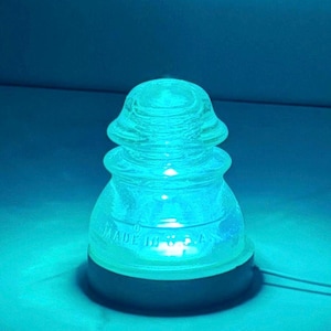 May include: A clear glass insulator lamp with a blue glow. The lamp has a rounded top and a base. The text "MADE IN U.S.A." is visible on the glass. The lamp is lit, casting a blue light.
