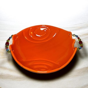 May include: An orange ceramic serving bowl with a glossy finish. The bowl features a spiral design in the center and beaded handles with red, green, and blue beads. The bowl is designed with a wavy edge.
