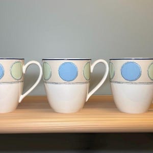 May include: Three ceramic mugs featuring a white base and a beige upper band decorated with alternating light blue and pale green circular patterns. Each mug has a curved handle and a thin dark blue rim along the top edge of the vessel.