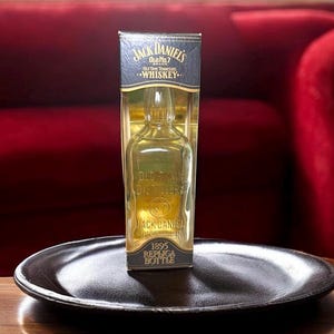 May include: A replica Jack Daniel's whiskey bottle in a clear plastic case. The bottle is empty and has a classic shape. The packaging features the Jack Daniel's logo and the text "Old No. 7 Brand Whiskey." The bottle is displayed on a dark, round tray.