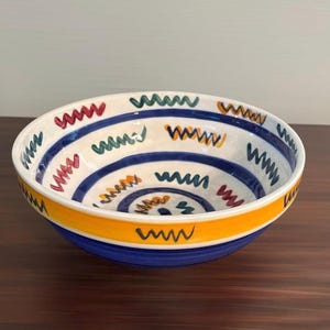 May include: A round ceramic bowl with a white interior and exterior featuring hand-painted blue horizontal stripes and scattered zigzag patterns in red, green, and yellow. The base of the bowl is solid blue with a wide yellow band above it.