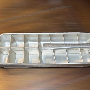 May include: A rectangular silver-colored metal ice cube tray with a built-in lever mechanism. The tray features a grid of dividers for creating ice cubes and includes a handle on one end. The metal lever is embossed with the text QUICKUBE.