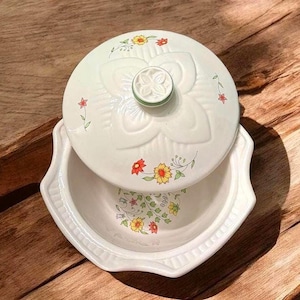 May include: A white ceramic serving dish with a lid. The lid and dish are decorated with a floral pattern in yellow, red, and green. The dish has a scalloped edge and sits on a wooden surface.