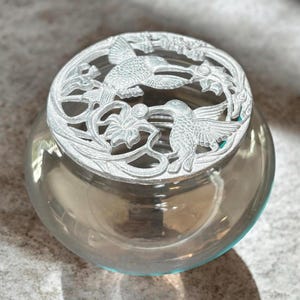 May include: A clear, rounded glass jar topped with a circular, silver-toned metal lid. The lid features an intricate, openwork design depicting two hummingbirds in flight, surrounded by detailed floral motifs and delicate, swirling vine patterns.