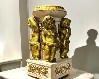 Puede incluir: Una escultura decorativa con tres figuras de querubines dorados, de pie alrededor de una columna blanca con una parte superior dorada. La base es blanca con detalles florales dorados. La escultura está sobre una superficie de mármol blanco.