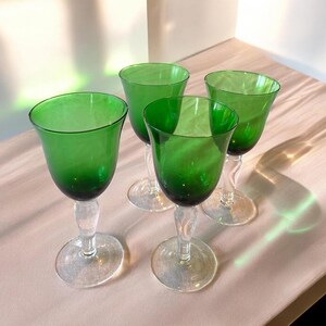 May include: Four green-tinted glass goblets with clear stems and bases. The goblets have a rounded bowl shape and are arranged on a light-colored surface. The glass has a vintage aesthetic.
