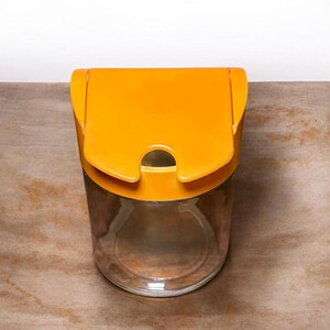May include: A clear glass storage jar featuring a vibrant mustard yellow plastic lid. The lid has a hinged design with a semi-circular cutout at the front edge, allowing for easy access to the contents inside the cylindrical glass container.