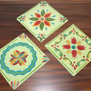 May include: Three square ceramic tiles featuring light green backgrounds and hand-painted floral motifs. The designs include red, orange, yellow, and blue petals arranged in symmetrical patterns with decorative borders and scalloped accents.