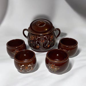 May include: A brown ceramic serving set with a lidded pot and four small bowls. The set features floral designs in white, orange, and gold. The pot has two handles. The bowls and pot have a gold rim.