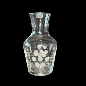 May include: A clear glass decanter with a wide base and a narrow neck. The decanter features an etched design of circular shapes and leaf patterns. A small green and white sticker is affixed to the top rim.