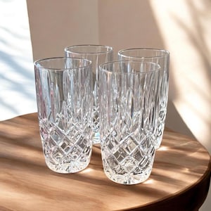 May include: Four clear, tall drinking glasses with a diamond-cut pattern. The glasses are made of crystal and sit on a wooden surface. The glasses are empty and ready to be filled with a beverage.