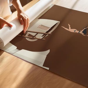 May include: A rolled-up print being unrolled, revealing a brown and beige illustration of a woman floating with an umbrella. The artwork features a ladder and architectural elements. The woman is wearing a bikini. The print is on a wooden surface.