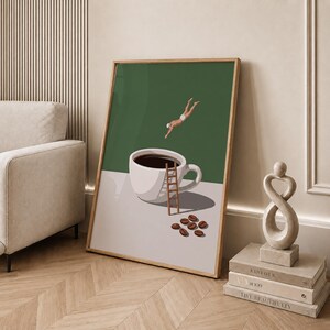 May include: A framed wall art print featuring a white coffee cup filled with dark liquid on a light surface. A small wooden ladder leans against the cup, while a figure in white swimwear dives towards it. Several brown coffee beans lie nearby.
