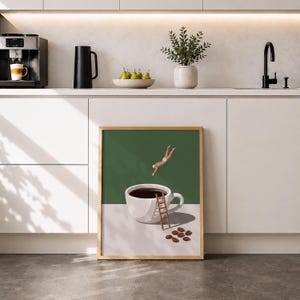 May include: A framed wall art print featuring a white coffee cup filled with dark liquid on a white surface against a green background. A small figure in white swimwear dives into the cup, while a wooden ladder leans against the side near coffee beans.