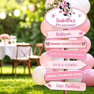 Pu&ograve; includere: Un cartello direzionale rosa e bianco per un ottavo compleanno, con Minnie Mouse. Il cartello indica agli ospiti i servizi igienici, il photo booth, la confetteria, i giochi, il castello gonfiabile e il face painting.