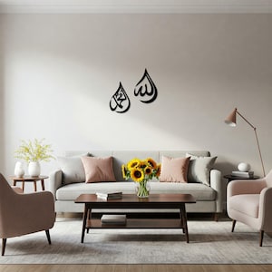 May include: Two black, stylized Arabic calligraphy wall art pieces. One piece features the name "Muhammad" and the other features the word "Allah". The art is mounted on a white wall above a gray sofa with pink pillows.