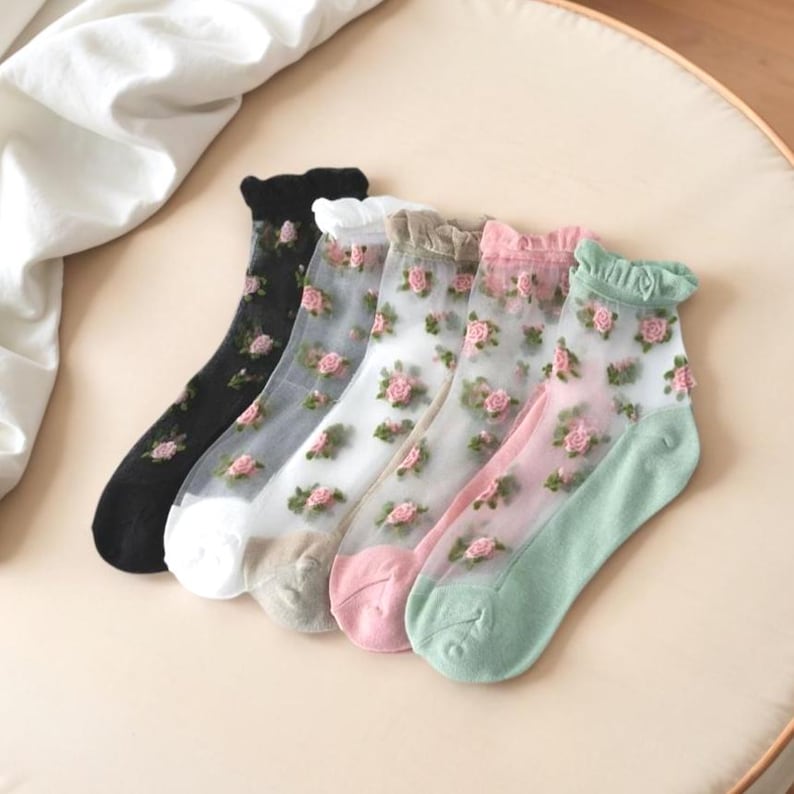 May include: Five pairs of sheer socks in various colors: black, white, tan, pink, and mint green. Each sock is adorned with embroidered pink roses and green leaves. The socks have ruffled tops and are arranged on a light-colored surface.