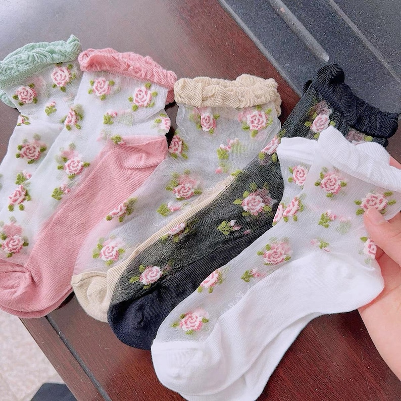 May include: Sheer socks in various colors, including white, pink, and black, adorned with embroidered pink roses and green leaves. Each sock has a ruffled trim at the top. The socks are lightweight and delicate, suitable for casual wear.