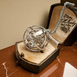 May include: A silver armillary sphere pendant necklace, presented in an open vintage jewellery box with a cream interior. The black box has gold trim, with the text "Jewellers" and "H. Moody" visible.