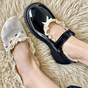 May include: Black patent leather Mary Jane shoes with a cream-colored lace trim and a small bow. The shoes are paired with white socks featuring floral embroidery and matching lace trim. The shoes have a thick sole.